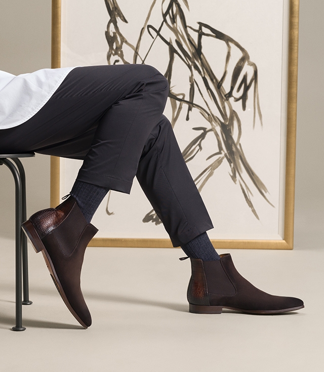 A male model sits in front of a painting styled in a white oxford and black pants wearing Magnanni’s Renley boots in brown.