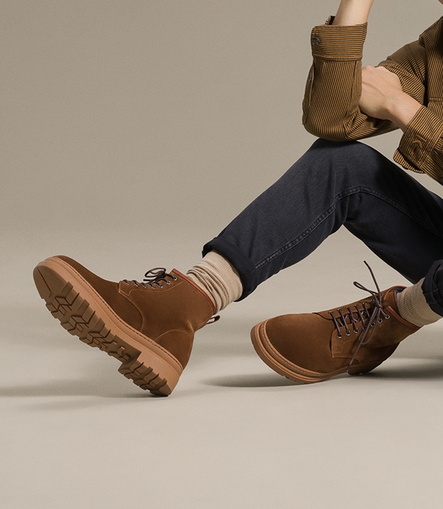 A male model seated on the ground styled in a brown shirt and navy jeans wears the Luther fashion boot in Cognac.