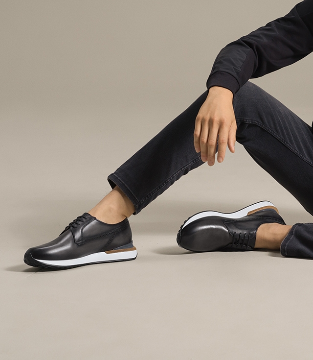 A male model wears the Leiva dress sneaker in Grey with black pants and black sweater. 