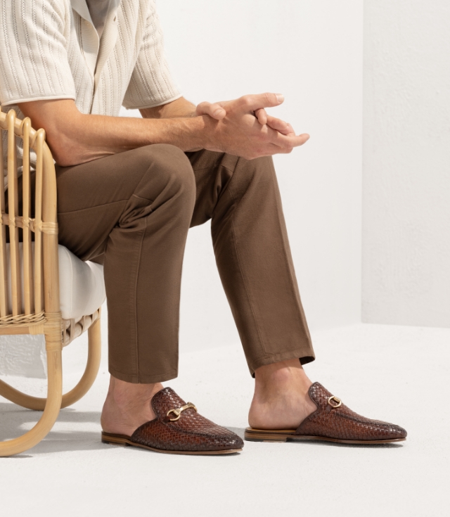 A man sits in a very short chair while wearing brown pants and Pachino Woven loafers.