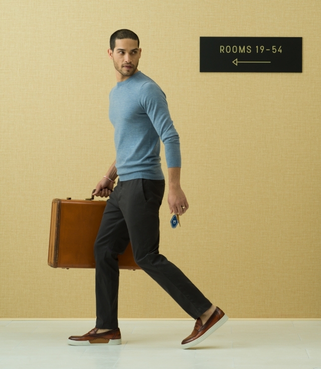 A man walks away looking to his left, holding a hotel key and suitcase while wearing a sky-blue shirt, black pants, and Magnanni Aydin Cognac loafers.