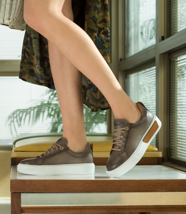 A woman stands on an end table while wearing Magnanni Amelia Taupe sneakers.