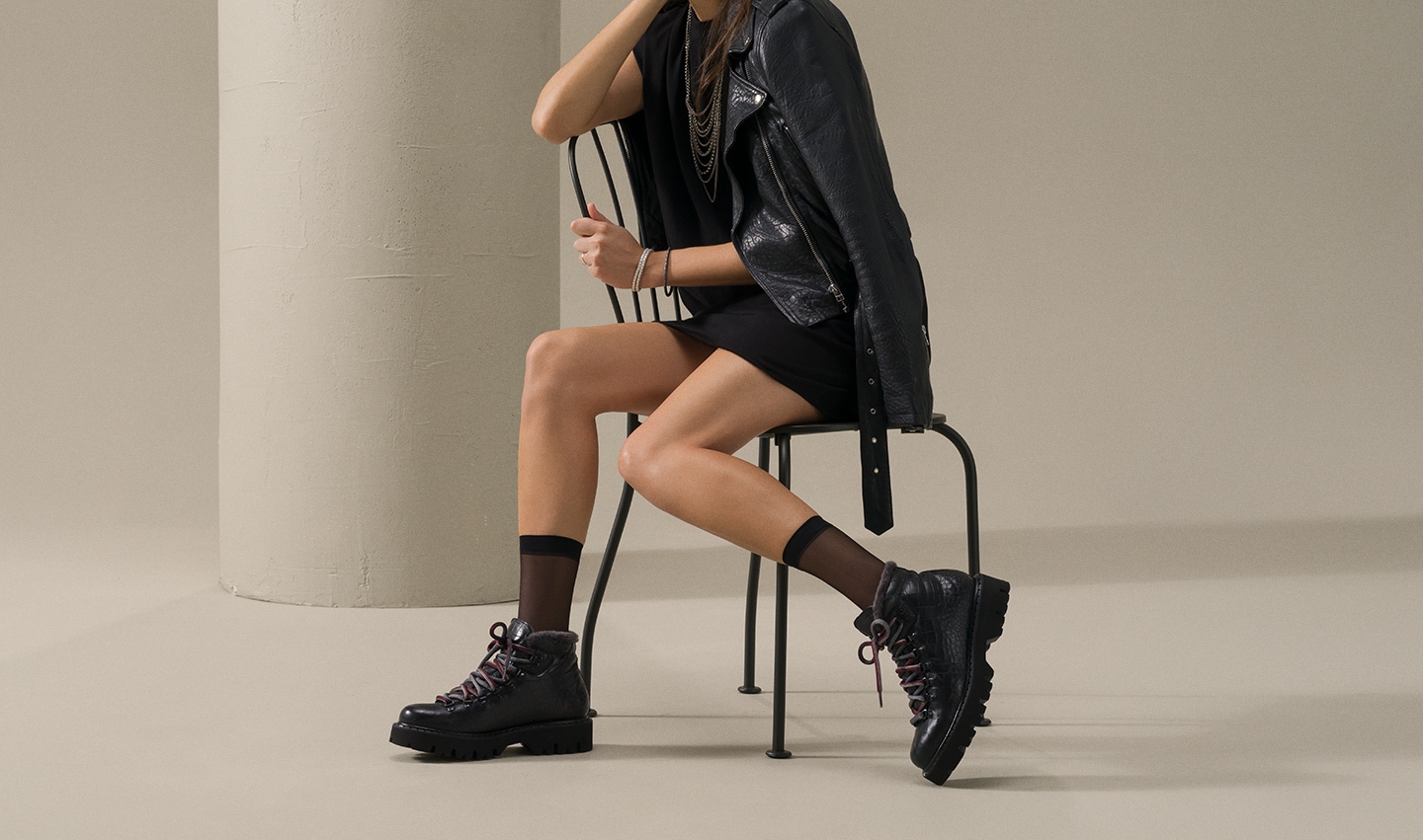 A woman sits in a metal chair while wearing a black dress and Magnanni Sierra boots in Black.