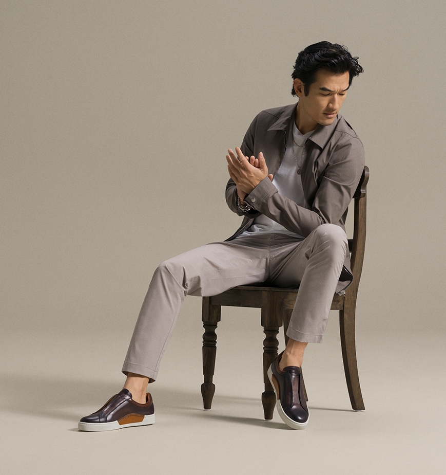 A male model sits in a wooden chair in light brown clothes and Magnanni Gasol sneakers.